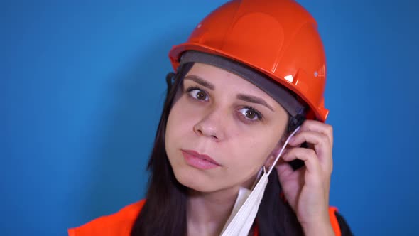 Female Construction Worker in Overalls Putting on Medical Mask on Face on Blue Background