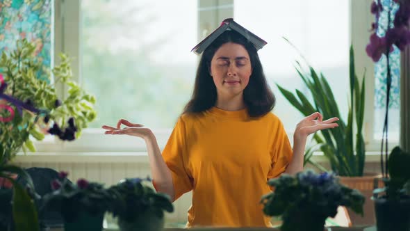 A young pretty woman holds book on her head and meditating with closed eyes.Indoor