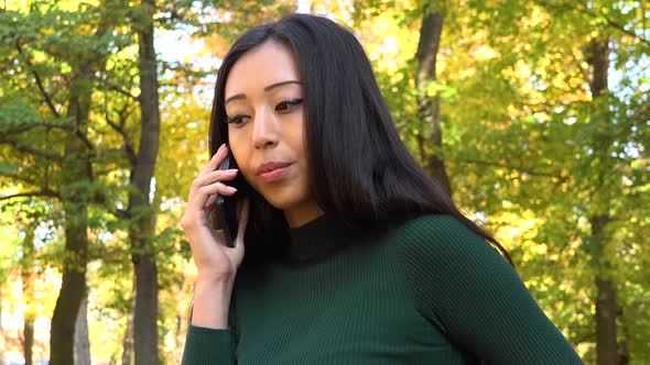 A Young Asian Woman Talks on A Smartphone in A Park