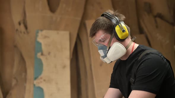 A carpenter wearing goggles, earmuffs and a respirator works in a workshop