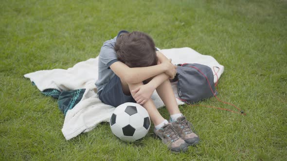 Top Angle View of Frustrated Caucasian Boy Sitting with Head on Knees, Pushing Away Soccer Ball, and