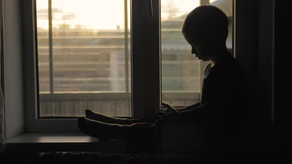 Silhouette Little Boy Uses Smartphone Sitting on Window Sill in His Room