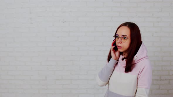 Young Woman in Glasses Speaks on Smartphone Standing Near White Brick Wall