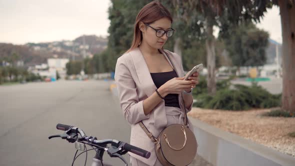 Young Brunette Woman Is Typing Sms on Mobile Phone Standing Near a Bike