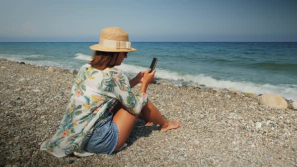 Attractive woman in straw hat sitting on beach, relaxing and using app on smartphone. 