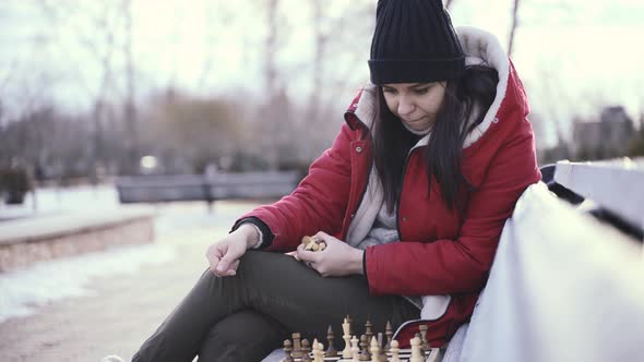 Young Woman in Winter Clothes Plays Chess Sitting on Bench in City Park