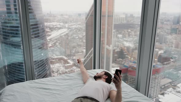 A Man Lying on a Bed with a Phone in His Hands in a Modern Apartment Near a Window Overlooking the