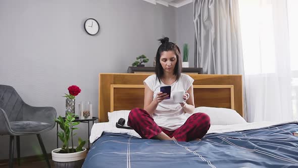 Brunette which Sitting on Bedroom Bed and Drinking Tea while Using Phone