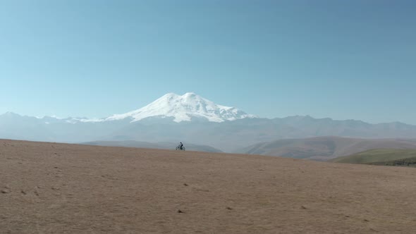 Person Rides Mtb Bicycle Along Rural Road in Field Against Distant Elbrus Mountain Under Clear Sky