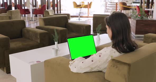 Young female entrepreneur using laptop with keyed Green Screen. She is sitting at hotel lobby chair