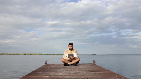 A guy with a tablet in his hands sits on a wooden pier near the river