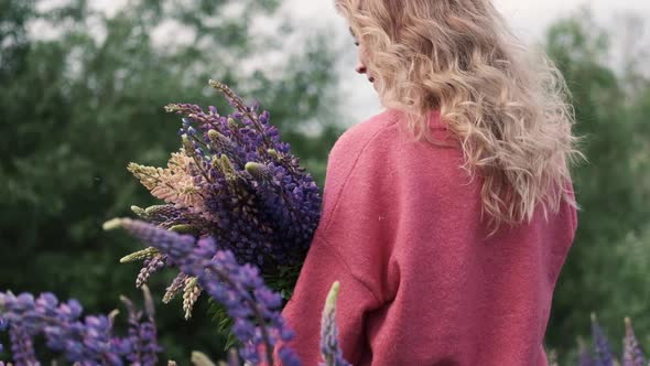 Young Beautiful Smiling Woman with a Bouquet Walks Among the Flowers of Purple Lupins