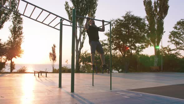 Fit Handsome Middle Aged Man with Long Gray Beard Doing Pullups on Horizon Bar on Sports Ground with