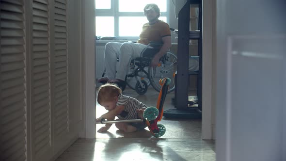 Father in a Wheelchair Watching His Little Baby Trying to Learn How to Skate on Scooter