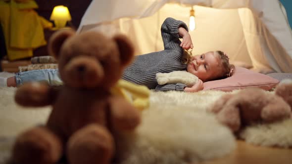 Wide Shot Relaxed Smiling Cute Girl Lying in Tent in Living Room with Blurred Teddy Bear at Front