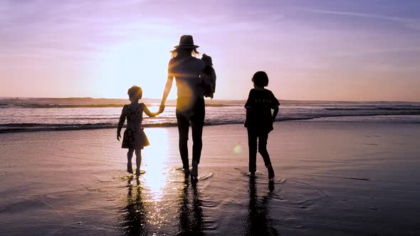 A mother holds hands and walks with her three kids at the beach in slow motion during a beautiful su