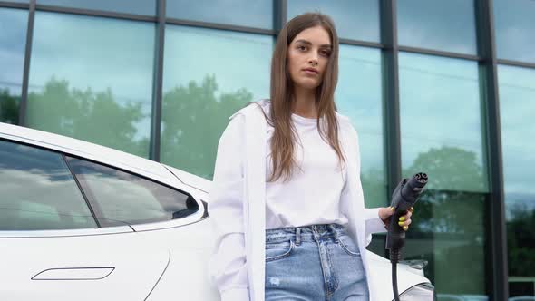 Smiling Young Caucasian Girl Plugging Electricity Cable in Electric Vehicle for Charging on Sunny