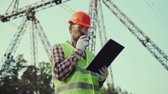 Caucasian Engineer in Protective Uniform and Orange Hard Hat Holds Walkietalkie and Clipboard on