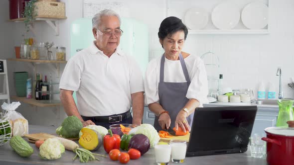 70s Elderly Senior Asian Couple in Relationship Using a Laptop While Cooking in Kitchen at Home