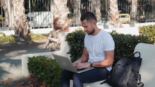 Smiling Young Man Working on the Laptop Sitting in Bench with Travel Case and Bag Backpack Tourist