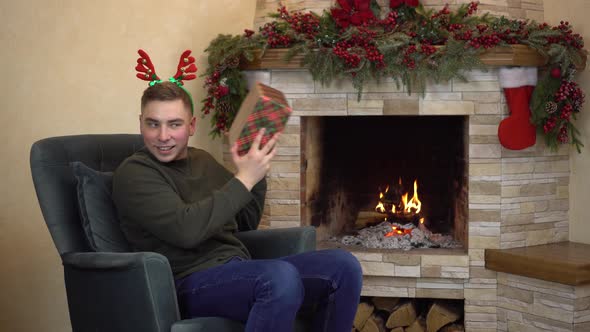 A Young Man Sits in an Armchair By the Fireplace with Horns on His Head and Holds and Shakes Gifts