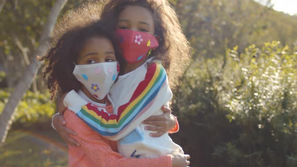 Happy Mixed Race Little Girls in Safety Mask Hugging and Smiling in Park