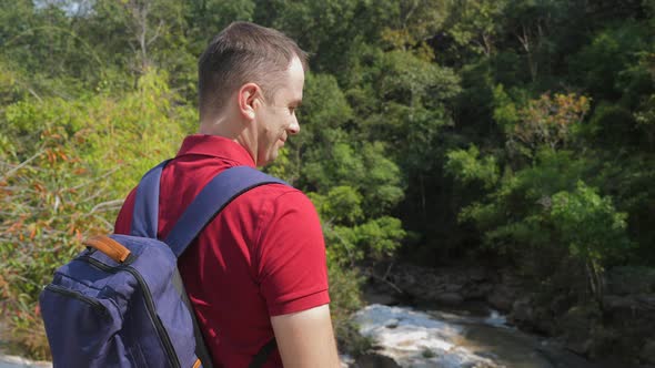 Travel Man in Red Polo Shirt with Backpack Standing Near Rock River
