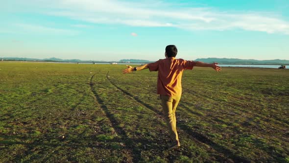 slow-motion of cheerful man running with arms raised across the green field with sunlight