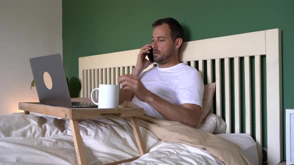 Businessman sitting in bed with laptop, talking on the phone