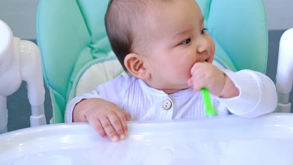 A hungry baby is gnawing on a plastic spoon at the table on a high chair. Teething, whims, itchy gum