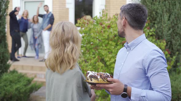 Pleasant Mature Couple with Cake Smiling and Looking Back at a Group of People Waving To Them