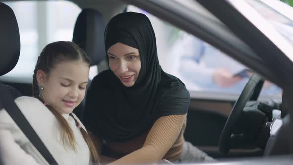 Positive Smiling Mother in Hijab Fastening Seat Belt for Daughter Sitting in Car