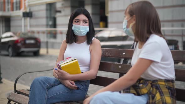 Portrait of Teenage Caucasian Girl with Black Hair in Coronavirus Face Mask Sitting on Bench on City