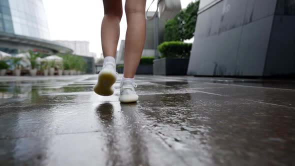 Female Feet in White Sneakers are Walking on the Wet Stone Slabs of a City Street