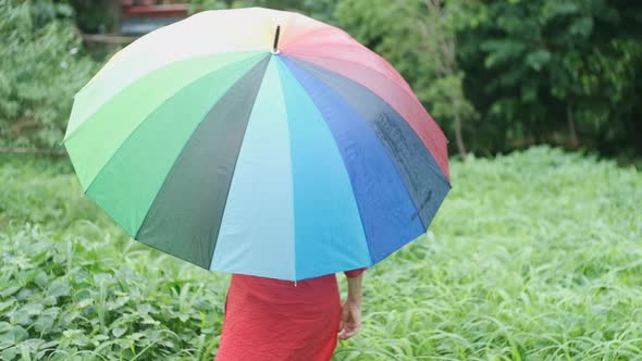 Pale skinned caucasian woman in red waling in garden in the rain with a rainbow umbrella slow motion