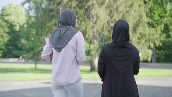 Back View of Positive Immigrants with Books Walking Outdoors on Sunny Day and Talking. Two Happy