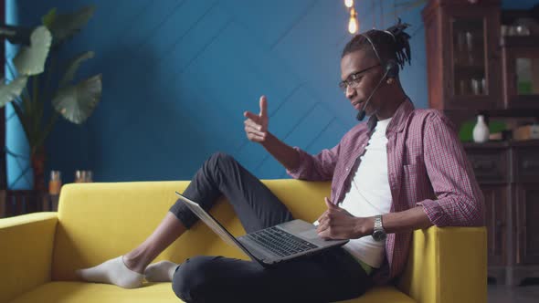Black Man Wearing a Headset is Sitting in Relaxed Position the Couch with Laptop and Talking on