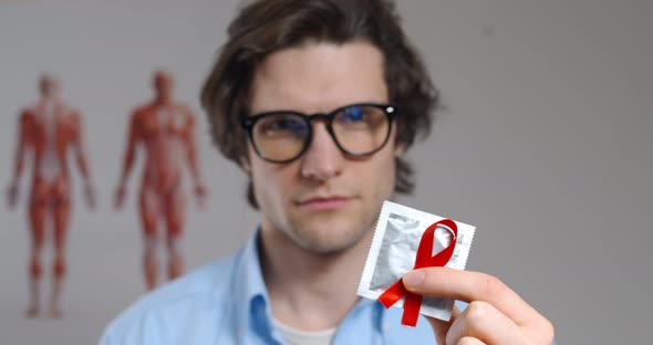 Close Up Portrait of Young Man in Glasses Holding Condom with Aids Awareness Red Ribbon