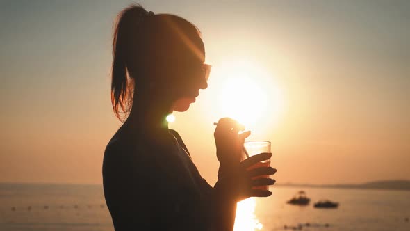 Silhouette Young Woman Drinking Cocktail on the Beach at Sunset on Background.