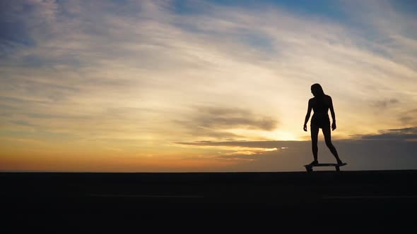 Silhouette of a Girl in Shorts and Sneakers Skateboarding Along the Road Against the Ocean and