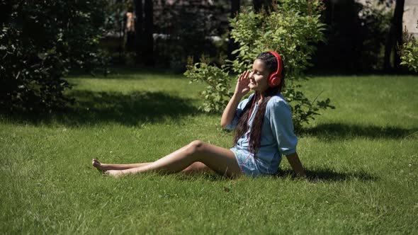 Young Girl Listening to Music on Headphones Using Smartphone Sitting on Grass in Park in Sunny
