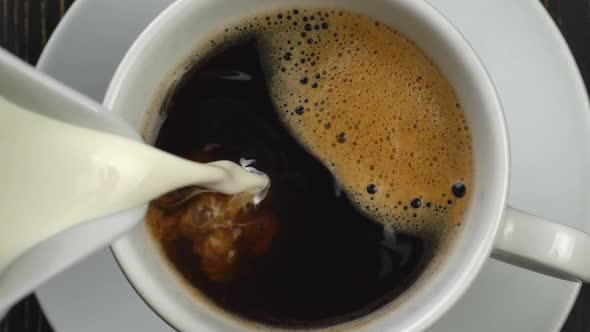 Tasty White Milk Slowly Pouring From a Jug Into a Cup of Brown Delicious Foamy Coffee. Top Down Slow