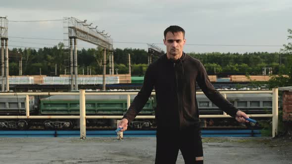 Adult man in sports clothes jumps on rope on roof against background of trains.