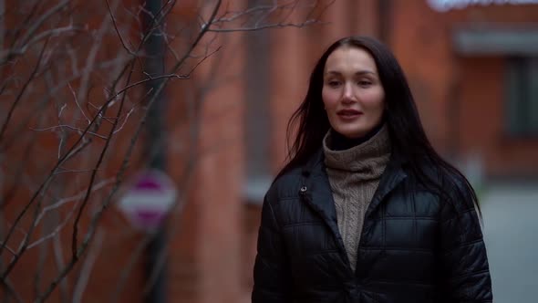 A Middle-aged Dark-haired Woman in a Black Jacket Walks on a City Street on an Autumn Day, Passing