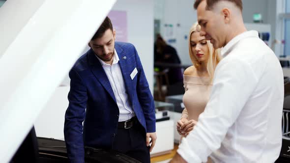 Salesman Showing Car Engine Under Open Hood to Couple in Auto Showroom Slow Motion