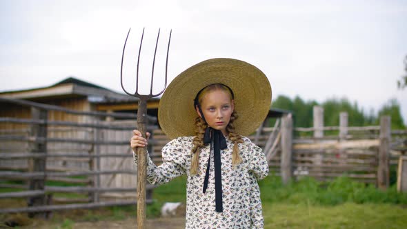 Serious Girl in Dress and Wicker Hat Posing with Hayfork in Village