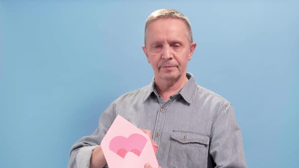 Aged Man Holding Open Pink Envelope with Pink Paper Heart on Blue Background