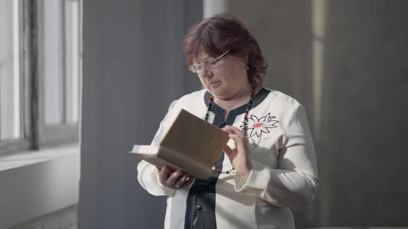 Intelligent Senior Caucasian Woman in Eyeglasses Reading Book Standing at Home in the Morning