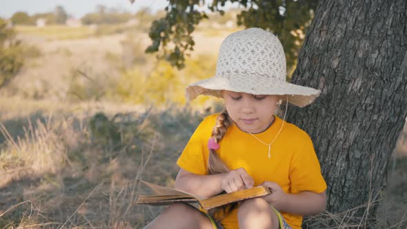 A girl is reading a book sitting under a tree. A child in a straw hat