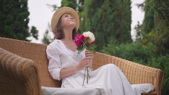 Satisfied Beautiful Woman in White Dress and Straw Hat Smiling Looking Up Admiring Bouquet of